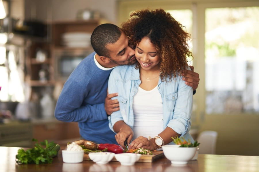 In a kitchen setting, a man embraces a woman who is cutting vegetables on a cutting board.