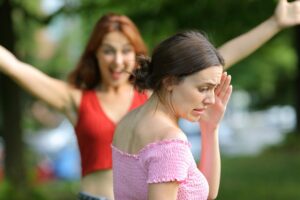 two women standing in the park, one with arms raised for an embrace while the other turns the face away.
