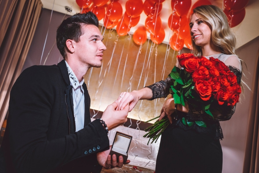 Man proposing, holding engagement ring, woman holding roses, red balloons in background.