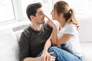 A woman playfully placing her finger on a guy's nose whule both sitting on a couch and looking at each other