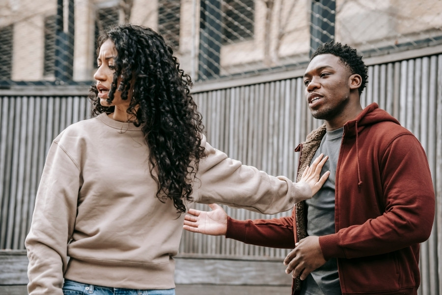 Woman extends arm defensively toward a man who has his hand on his chest.