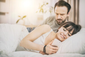 A couple on the bed with the woman facing the other side while checking her phone but the man appears to wonder what she is checking.