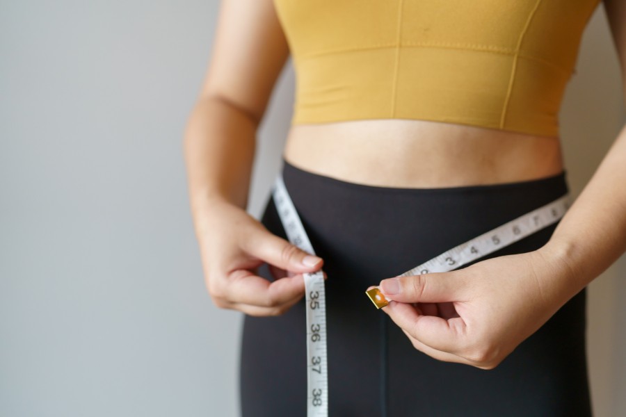 Torso view of a woman in exercise attire measuring her waist with a tape measure.
