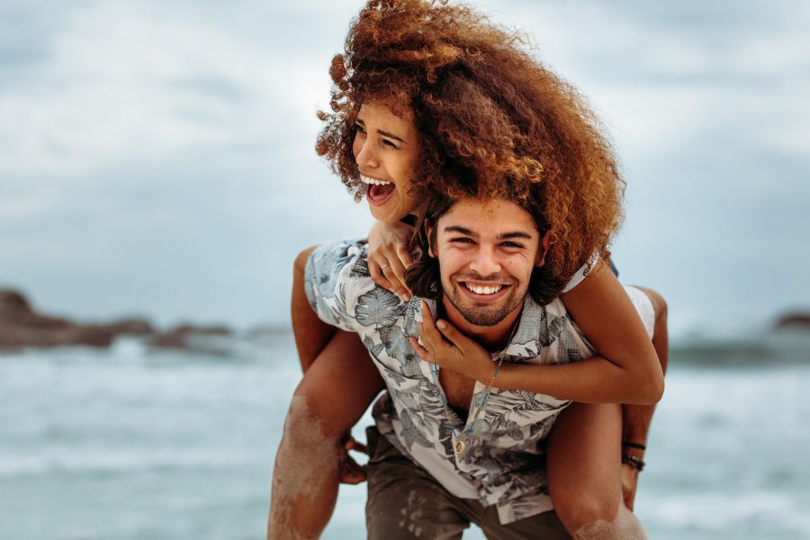 Romantic couple on a beach; a woman with curly hair is on a man's back in a piggyback ride, both are smiling.