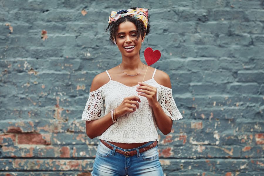 A smiling woman with a headscarf, off-the-shoulder white blouse, and denim shorts stands in front of a weathered brick wall while holding a red heart on a stick.
