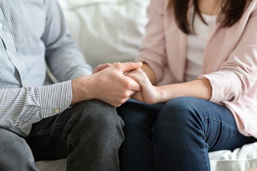 A man and a woman sit closely, holding hands in a gesture of comfort and connection.