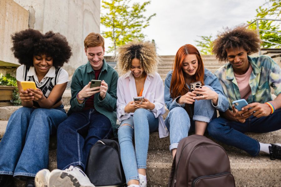 Five young adults sit on outdoor steps, each looking at their mobile phones.