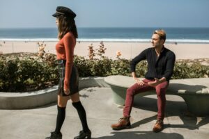 A lady walking away from a man that is sitting on a bench near the beach