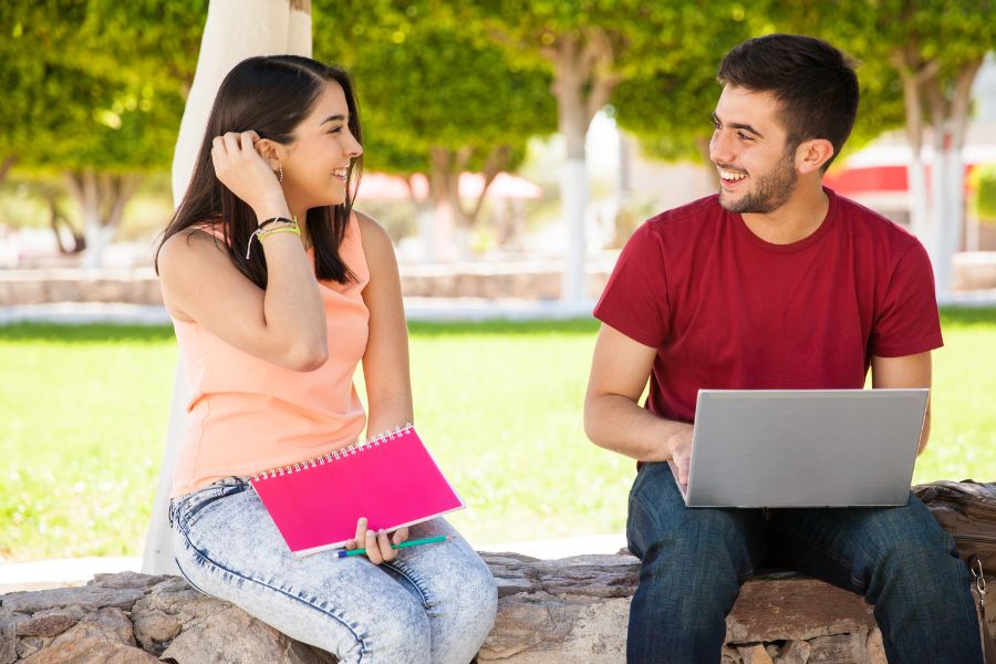 A young man and woman sitting outside on a low rock wall looking at each other and smiling; he is holding a laptop and she is holding a notebook and pencil.