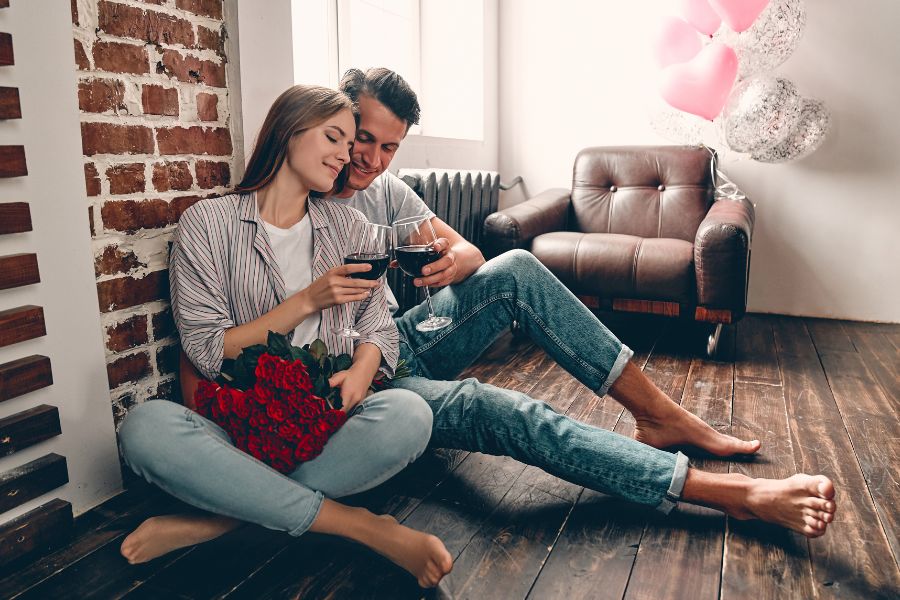 Couple sitting barefoot on a wooden floor with wine and red roses.