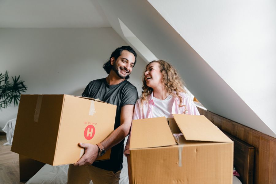 A smiling couple carries cardboard boxes in a room with slanted ceilings.