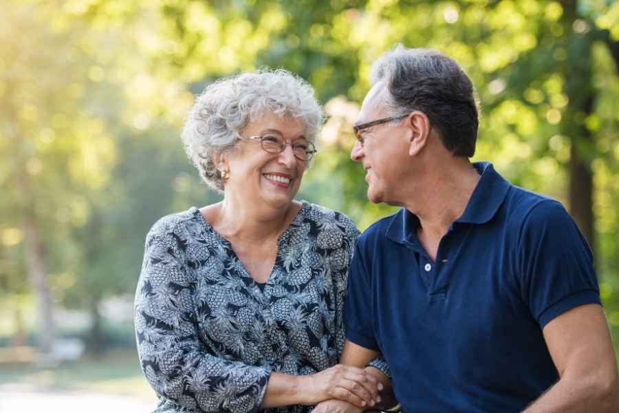 Elderly couple looking at each other, smiling, while holding hands outdoors.